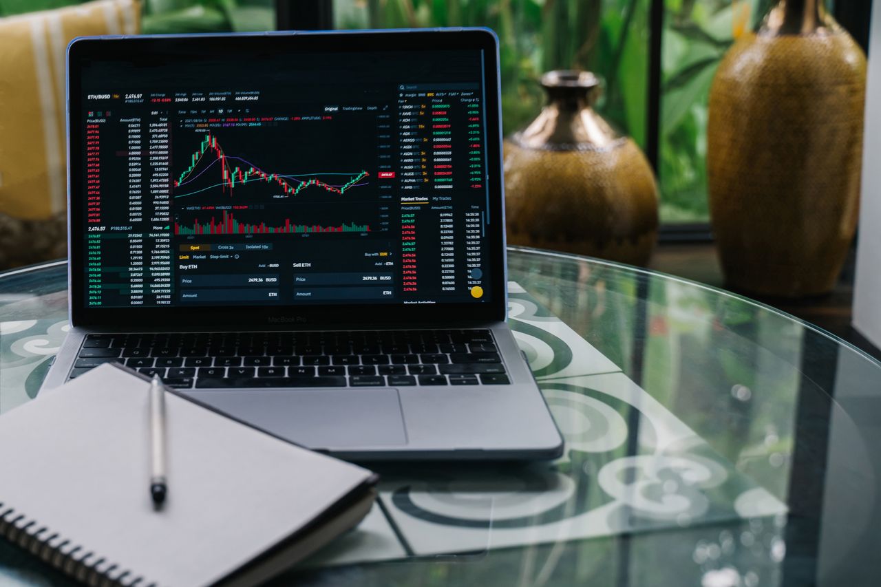 home-services-004 Laptop displaying cryptocurrency stocks and graphs on a glass table with a notepad.
